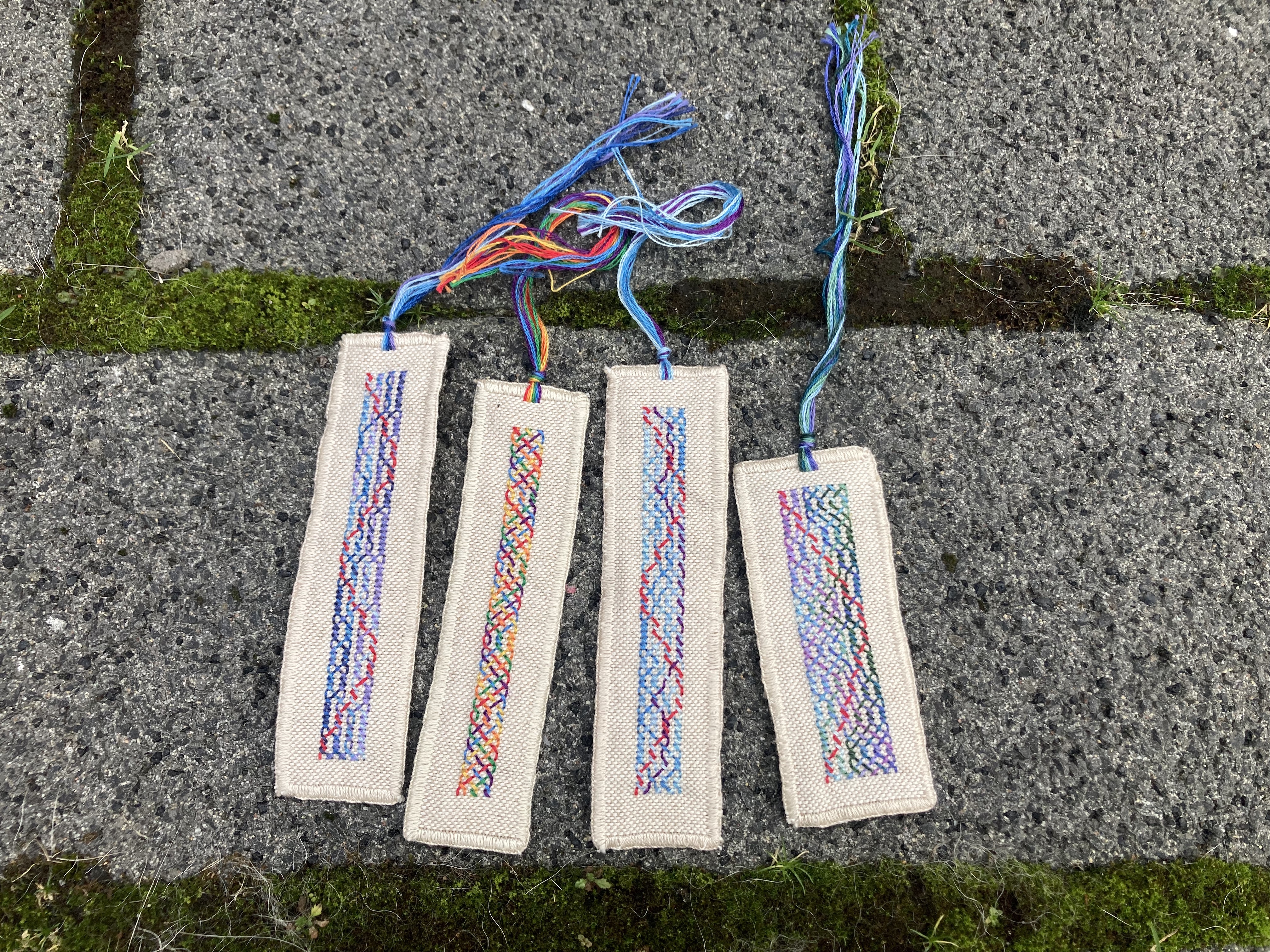 Four embroidered bookmarks with colorful tassels lying on gray bricks with bits of moss between the bricks
